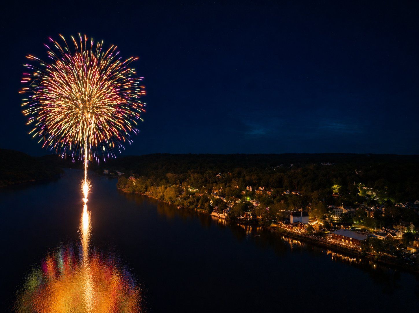 Fireworks bursting over the Delaware River beside New Hope at night, the town lit up along the bank